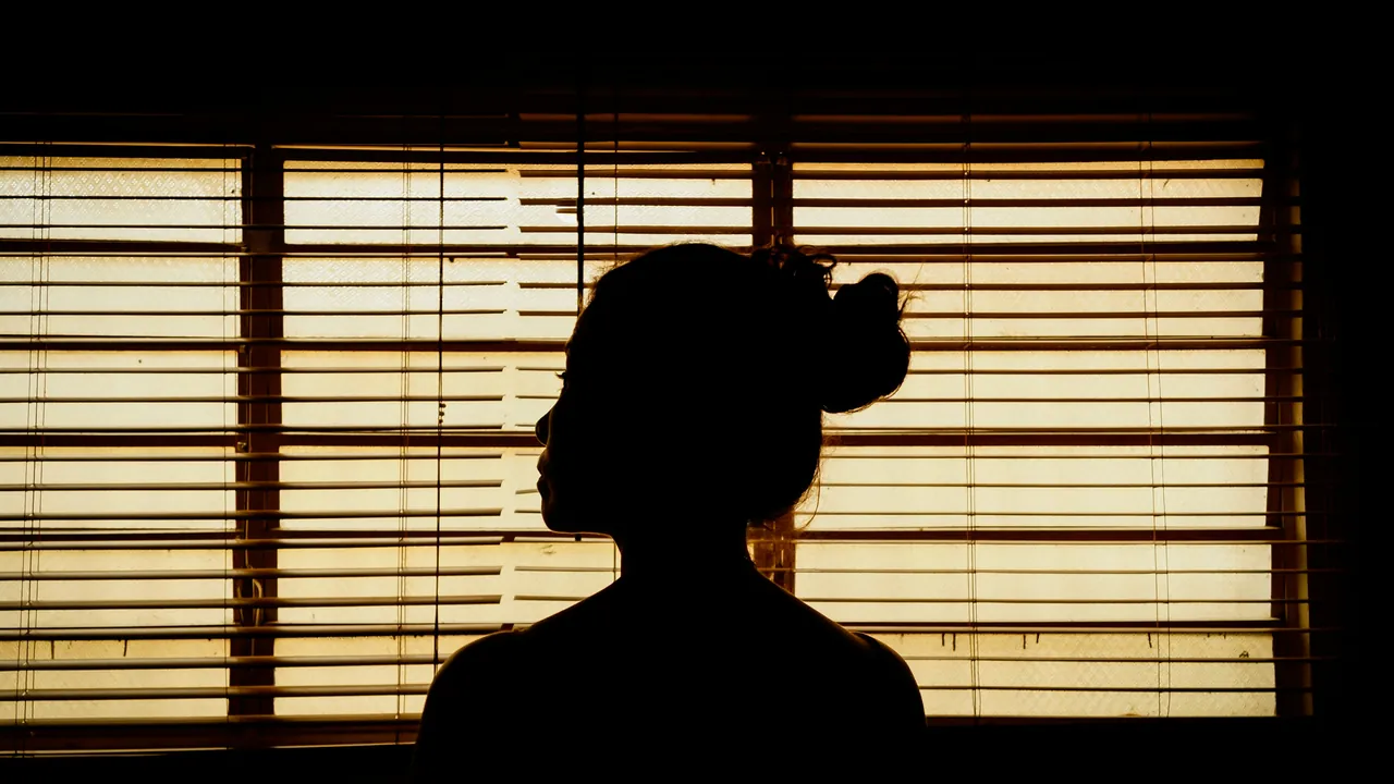 A silhouette in front of lit window blinds, warm light through the slats.