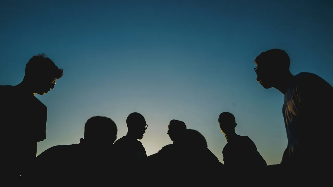 A group in silhouette against a deep blue evening sky, leaning in to talk.
