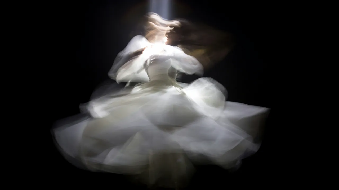 A dancer in a white dress, blurred into a spinning cloud against black.