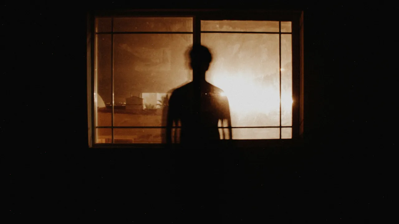 A figure in silhouette at a glowing window in a dark room.